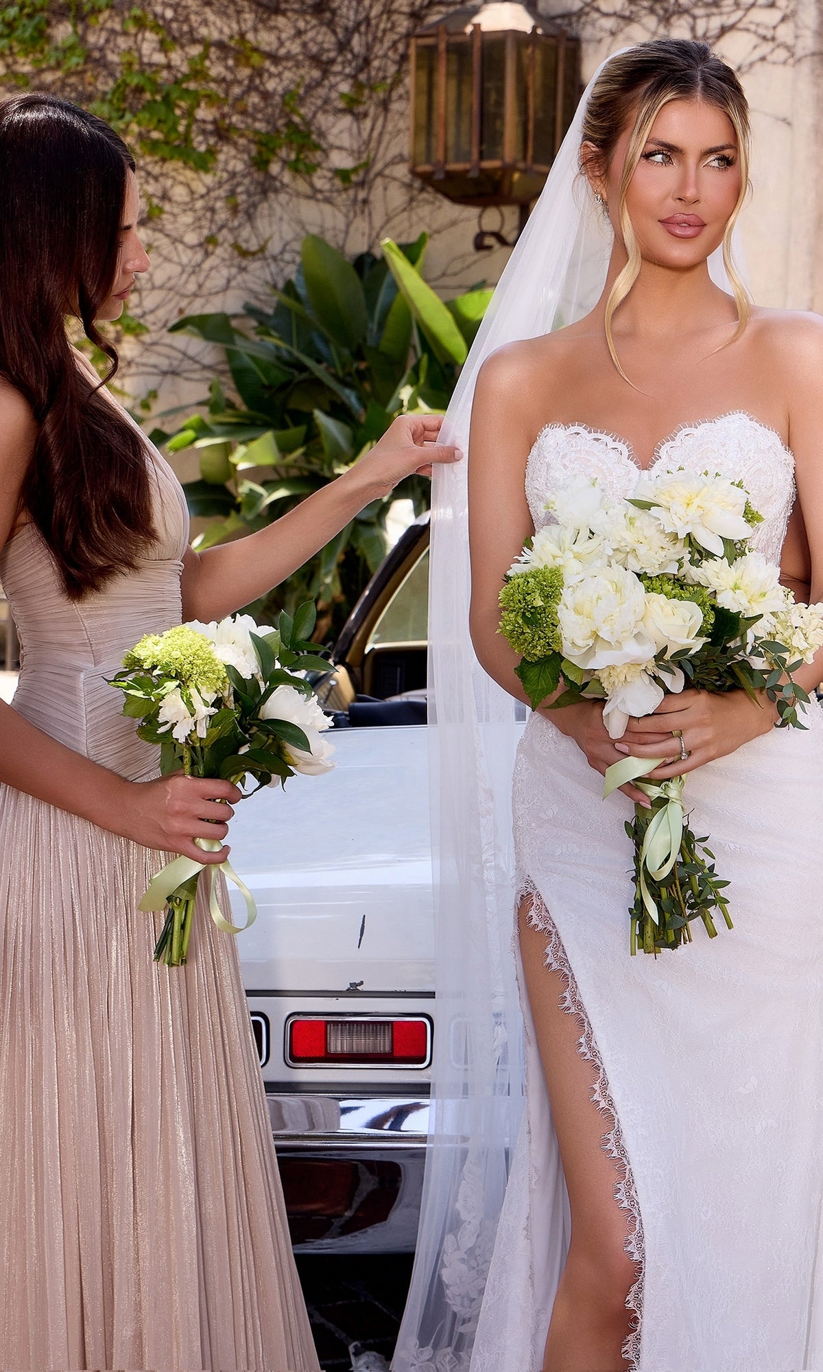 A bride wearing the Ladivine CDS550W long wedding dress holds a bouquet while a bridesmaid in a beige dress adjusts her veil. Both are standing beside a white car with greenery in the background.